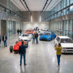 Des visiteurs photographient une collection de voitures de course légendaires dans un garage moderne et lumineux au Mans.